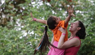 A woman joyfully lifts a smiling girl with a long ponytail and colourful ribbons into the air outdoors, both reaching their arms upward against a background of green trees.