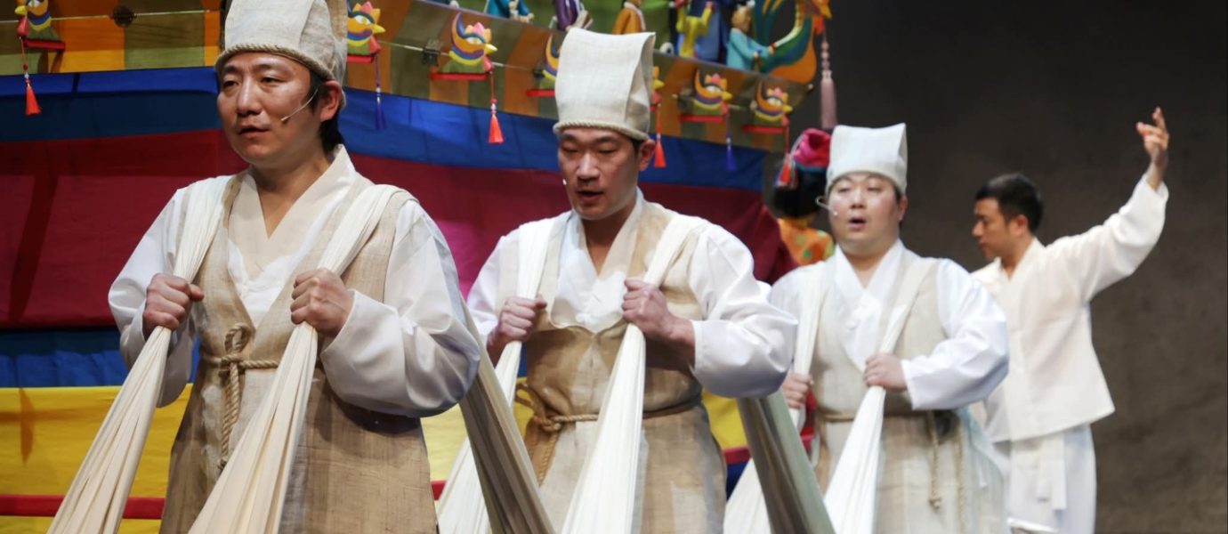 Three people in traditional Korean attire pull long white fabric in front of a colourful, decorated float; a fourth person stands behind them with one arm raised, partially blurred.