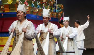 Three people in traditional Korean attire pull long white fabric in front of a colourful, decorated float; a fourth person stands behind them with one arm raised, partially blurred.