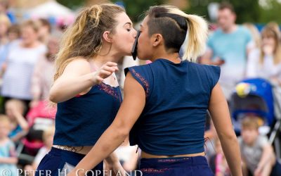 Two women wearing matching navy blue outfits perform closely, touching faces with a black object between their mouths, in front of a blurred crowd at an outdoor event.
