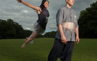 A barefoot man stands on grass, looking ahead, whilst a barefoot woman in a black dress leaps joyfully behind him with arms outstretched. The sky is cloudy, and they are outdoors in a park with trees in the background.
