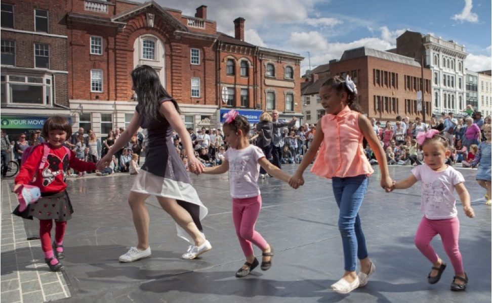 A group of children and an adult hold hands and dance in a circle outdoors on a city square, with people watching and historic buildings in the background under a partly cloudy sky.