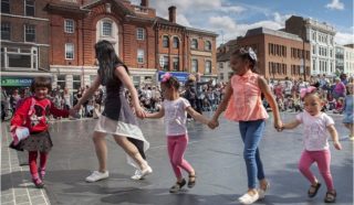 A group of young girls and a woman hold hands and dance in a circle outdoors, with a crowd and historic buildings in the background under a partly cloudy sky.