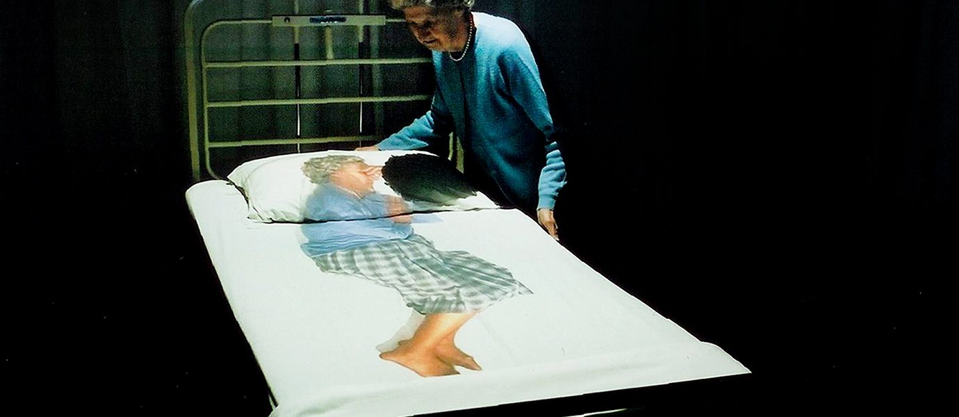 An elderly woman stands by a hospital bed, looking at bedcovers printed with a life-sized image of herself curled up, suggesting themes of memory, ageing, or vulnerability.