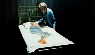 An elderly woman stands by a hospital bed, looking at bedcovers printed with a life-sized image of herself curled up, suggesting themes of memory, ageing, or vulnerability.
