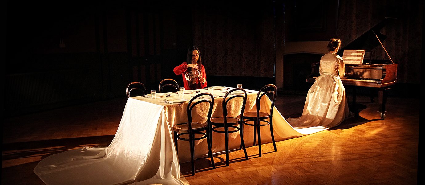 Two women in elegant gowns sit in a dimly lit room; one plays a grand piano whilst the other sits at a long, white-draped table set for dinner, illuminated by warm light.
