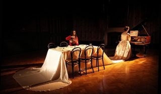 Two women in elegant gowns sit in a dimly lit room; one plays a grand piano whilst the other sits at a long, white-draped table set for dinner, illuminated by warm light.