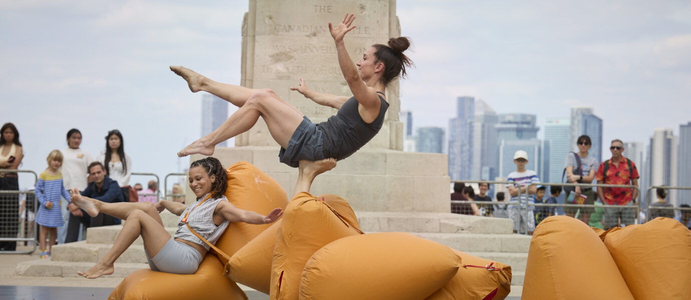Two women perform an outdoor dance, leaping onto large orange cushions. One woman is mid-air, arms and legs extended. The background shows a monument, onlookers, and a city skyline under a cloudy sky.