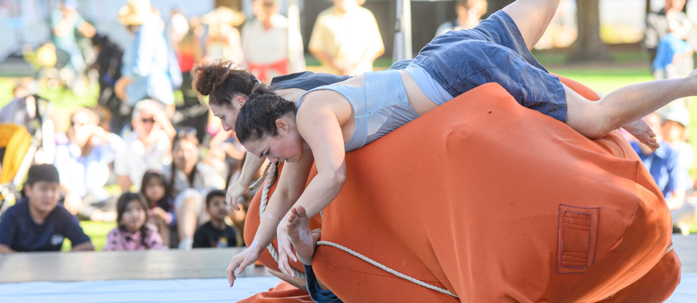 Two people tumble and wrestle on top of a large orange cushion outdoors, with a crowd of spectators watching in the background.