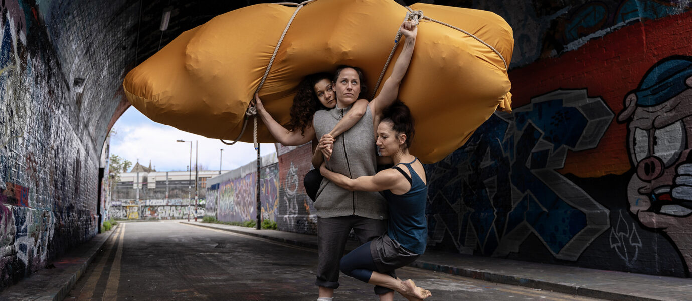 Three people stand and balance under a large, yellow, inflated shape in a graffiti-covered tunnel. One person is suspended off the ground whilst clinging to the others. The background shows an urban street scene.