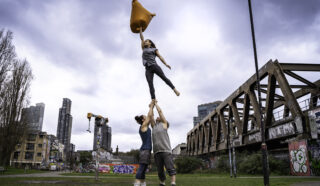 Three people perform acrobatics outdoors; two lift a woman holding an orange cushion high in the air. Graffiti covers a bridge nearby; city buildings and cloudy sky are in the background.