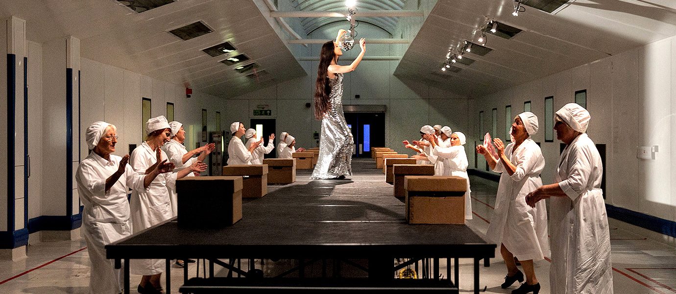 A woman in a silver dress walks a catwalk in a factory, while workers in white uniforms and caps stand on either side, clapping and smiling.