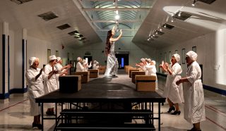 A woman in a silver dress walks a catwalk in a factory, while workers in white uniforms and caps stand on either side, clapping and smiling.