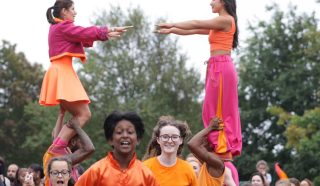 A lively group performs outdoors: two duos form human towers whilst others in orange and pink costumes dance and smile, with a cheerful crowd watching in the background.