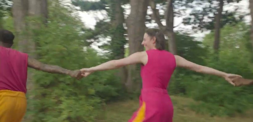 Two people, one in a bright pink dress and one in a pink top with yellow trousers, hold hands and dance in a circle outdoors among trees and greenery.