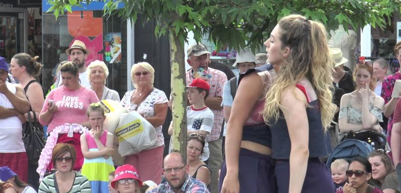 Two women stand closely together under a tree, performing outdoors for a diverse crowd. People of all ages watch, some seated and some standing, on a sunny day in a busy public area.
