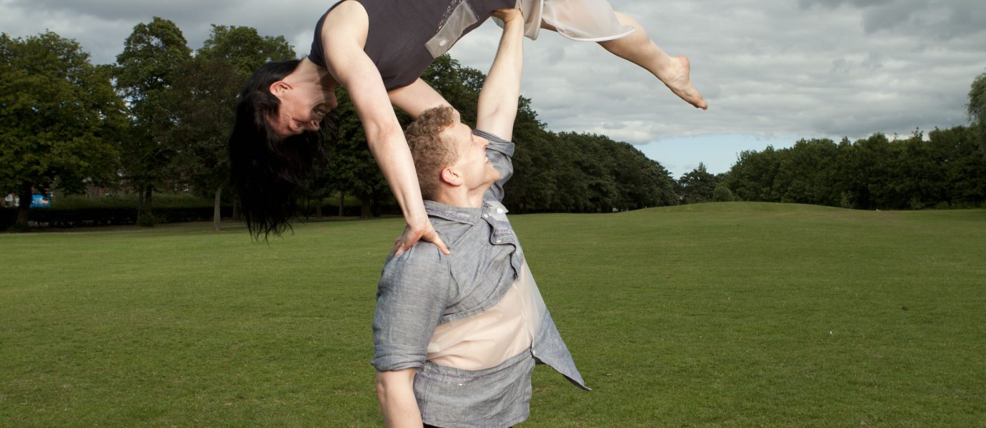 A barefoot man stands on grass, balancing a barefoot woman upside down above his head with one hand. They are both outdoors in an open field under a cloudy sky.