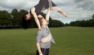 A barefoot man stands on grass, balancing a barefoot woman upside down above his head with one hand. They are both outdoors in an open field under a cloudy sky.