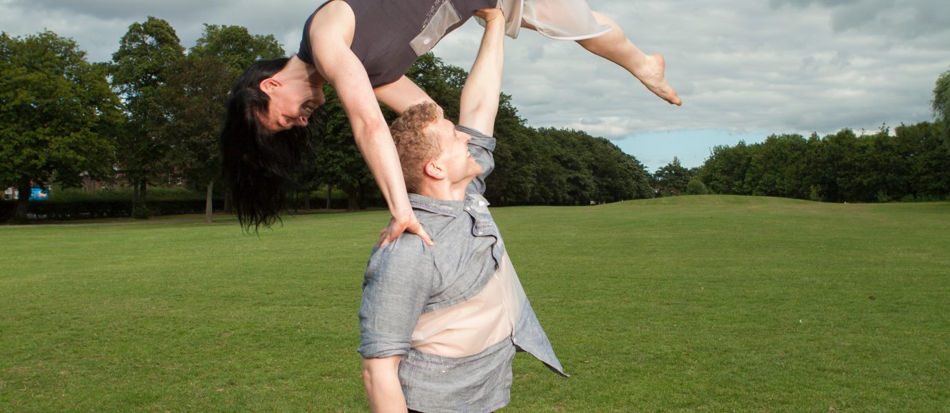 A man balances a woman above his head with one arm in a grassy park. The woman is upside down, legs extended, while the man stands barefoot, both wearing casual clothing. Trees and cloudy sky are in the background.