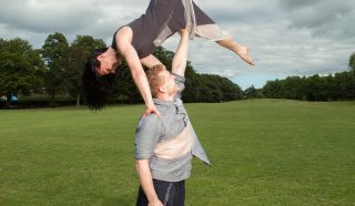 A man balances a woman above his head with one arm in a grassy park. The woman is upside down, legs extended, while the man stands barefoot, both wearing casual clothing. Trees and cloudy sky are in the background.