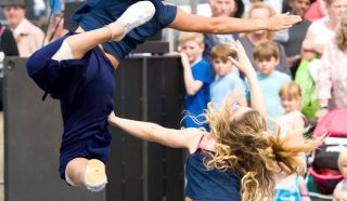 Two dancers perform energetically outdoors; one leaps mid-air with extended limbs while the other kneels, reaching up. A crowd of onlookers, including children, watches in the background.