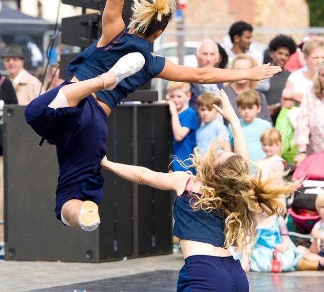 Two dancers perform energetically outdoors; one leaps mid-air with extended limbs while the other kneels, reaching up. A crowd of onlookers, including children, watches in the background.