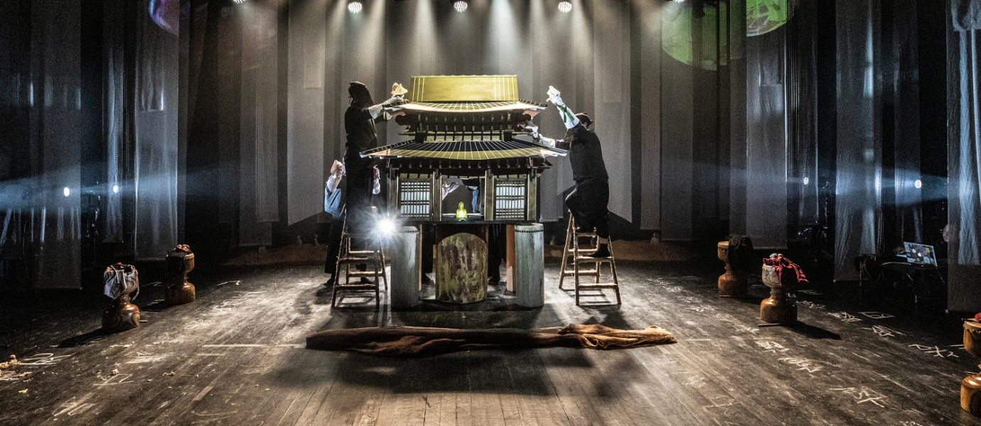 Four people dressed in black stand on ladders around a detailed model building on a stage, illuminated by dramatic overhead lighting with props and curtains in the background.