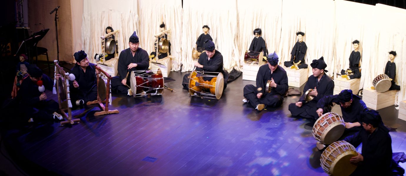 A group of musicians in traditional black attire play drums and gongs on a stage, with a row of white-robed figures seated behind them, possibly traditional puppets or mannequins.