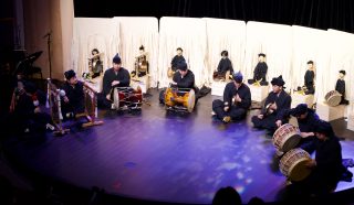 A group of musicians in traditional black attire play drums and gongs on a stage, with a row of white-robed figures seated behind them, possibly traditional puppets or mannequins.