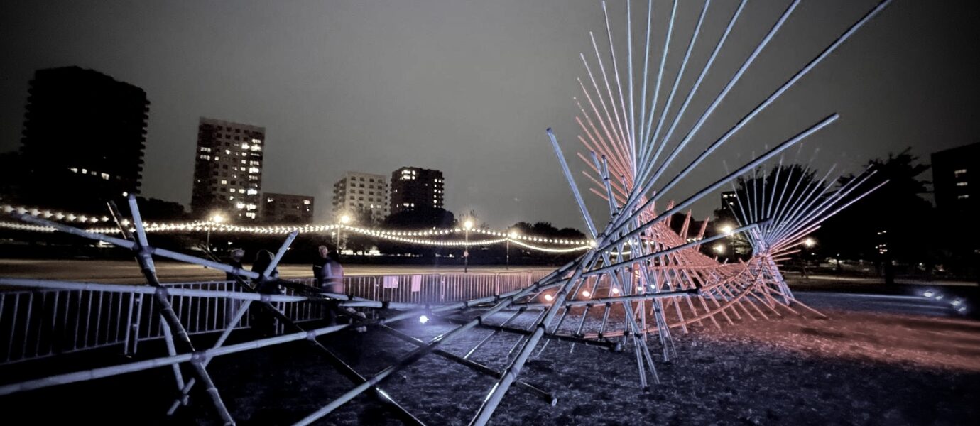 A nighttime photo shows a modern outdoor sculpture with intersecting metal rods lit by coloured lights, set against a cityscape with tall buildings and fairy lights on a bridge in the background.