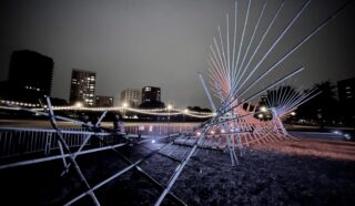 A nighttime photo shows a modern outdoor sculpture with intersecting metal rods lit by coloured lights, set against a cityscape with tall buildings and fairy lights on a bridge in the background.