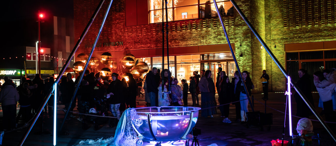 A crowd gathers at night around a large illuminated metal bowl with light tubes, in front of a modern brick building. Colourful lights highlight the scene.
