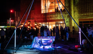 A crowd gathers at night around a large illuminated metal bowl with light tubes, in front of a modern brick building. Colourful lights highlight the scene.