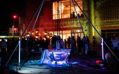 A crowd gathers at night around a large illuminated metal bowl with light tubes, in front of a modern brick building. Colourful lights highlight the scene.