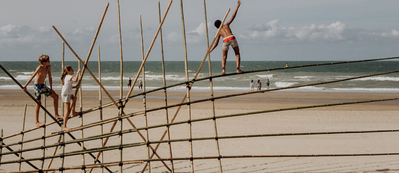Three children climb and balance on a bamboo structure on a sandy beach, with the sea and a few people walking in the background under a partly cloudy sky.