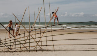Three children climb and balance on a bamboo structure on a sandy beach, with the sea and a few people walking in the background under a partly cloudy sky.