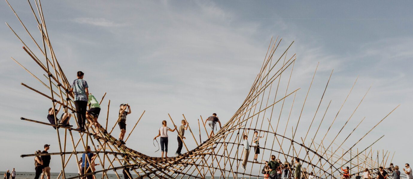 People climb and walk on a large bamboo art installation shaped like a wave on a sandy beach under a clear blue sky.