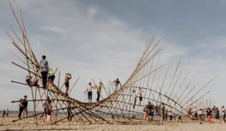 People climb and walk on a large bamboo art installation shaped like a wave on a sandy beach under a clear blue sky.