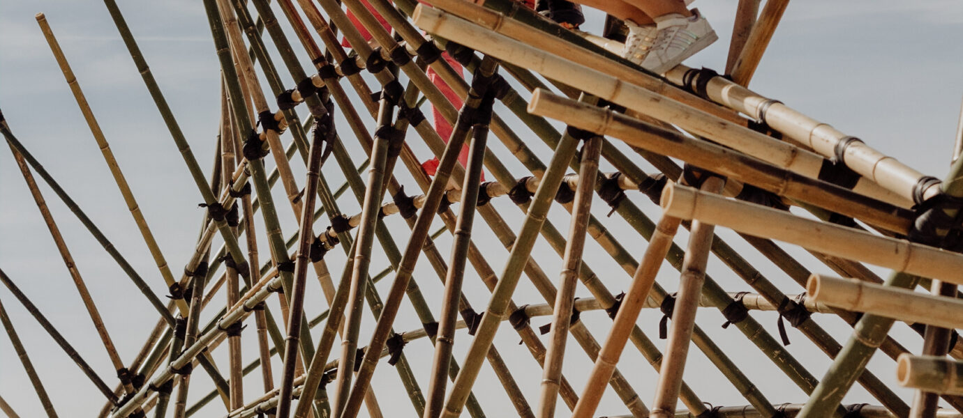 Four children climb and play on a large bamboo structure on a sandy beach under a clear sky, appearing adventurous and excited.