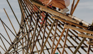 Four children climb and play on a large bamboo structure on a sandy beach under a clear sky, appearing adventurous and excited.