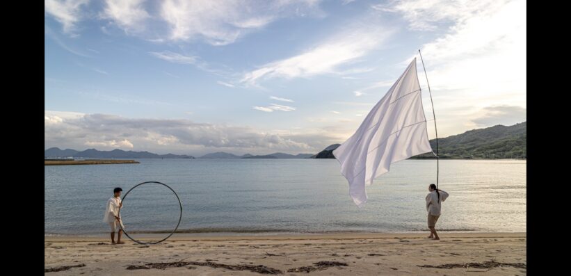 Two people walk along a sandy beach; one carries a large white flag, and the other holds a large metal ring. The calm sea and distant mountains are visible under a partly cloudy sky.