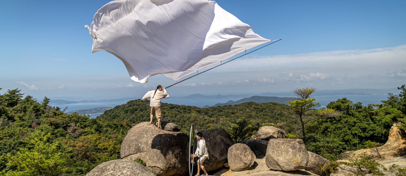 Two people stand on large rocks atop a hill, one holding a tall pole with a large white flag billowing dramatically in the wind, surrounded by green trees and distant mountains under a blue sky.