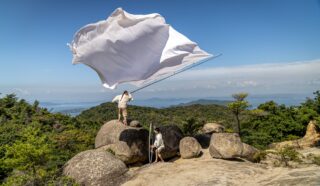 Two people stand on large rocks atop a hill, one holding a tall pole with a large white flag billowing dramatically in the wind, surrounded by green trees and distant mountains under a blue sky.