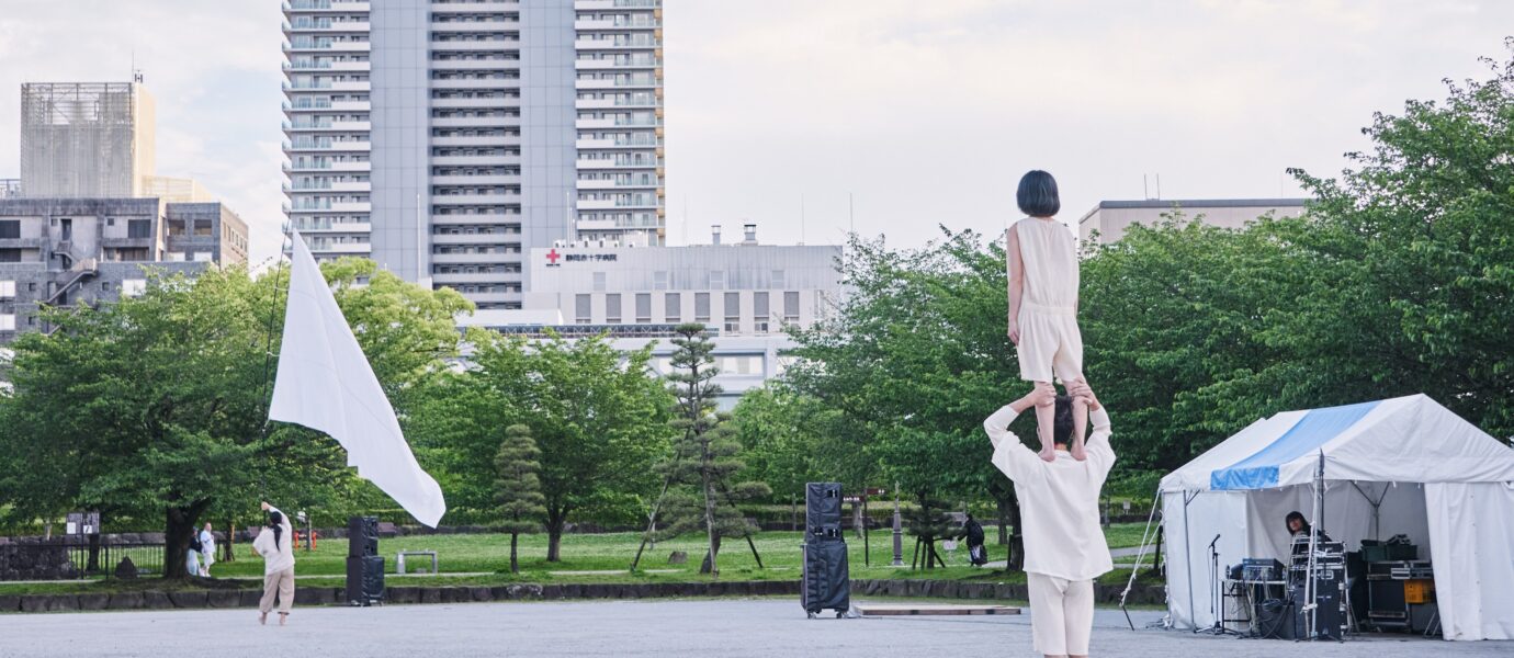 A person stands on another's shoulders in a park, while someone with a large white flag walks nearby. Buildings, green trees, a white marquee, and audio equipment are visible in the background under a partly cloudy sky.