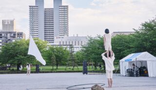 A person stands on another's shoulders in a park, while someone with a large white flag walks nearby. Buildings, green trees, a white marquee, and audio equipment are visible in the background under a partly cloudy sky.