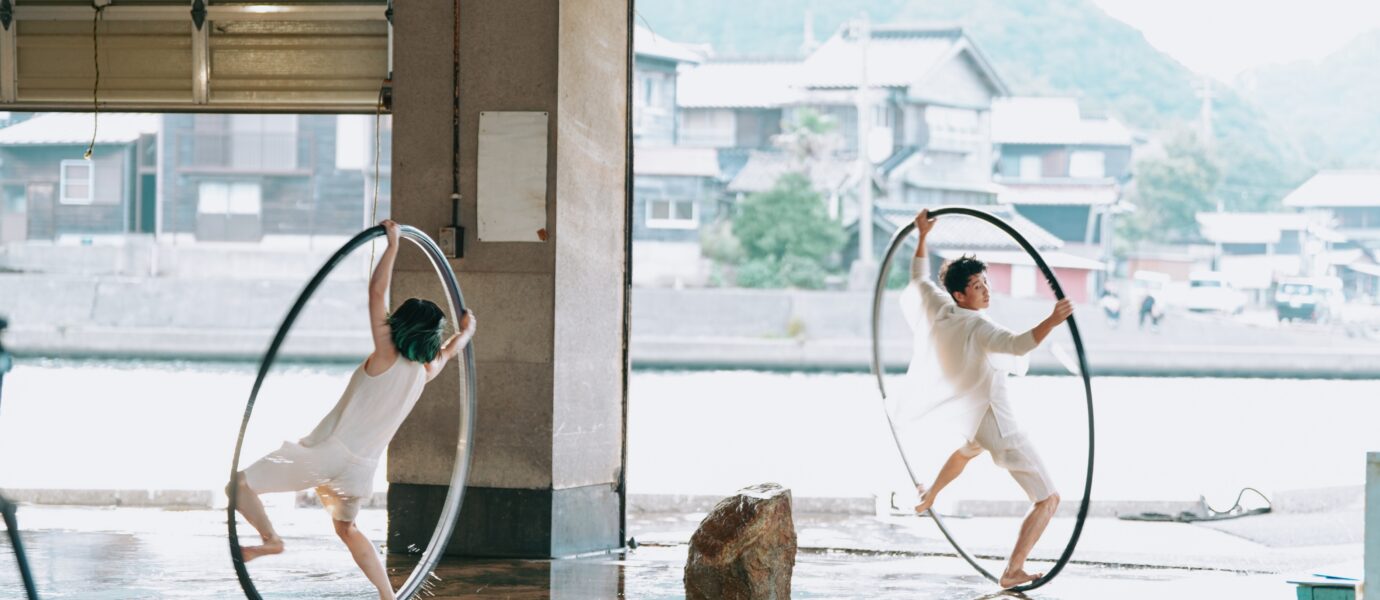 Two performers in white outfits balance inside large metal hoops on a wet floor, with houses and mountains visible across the water in the background.
