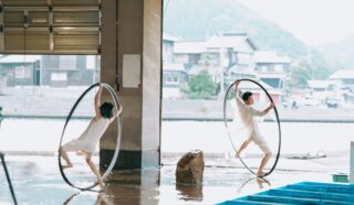 Two performers in white outfits balance inside large metal hoops on a wet floor, with houses and mountains visible across the water in the background.