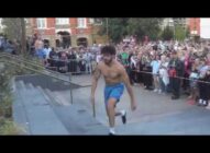 A bare-chested man in blue shorts runs up steps in an outdoor urban area, as a large crowd watches behind a barrier.