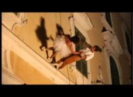 Two dancers perform attached to the side of a building at night, appearing to defy gravity as they move horizontally, with their shadows cast on the wall behind them.