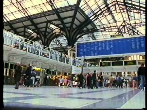 Busy railway station interior with arched glass roof, a large blue digital departures board, and many people walking or waiting. Natural light fills the spacious concourse.